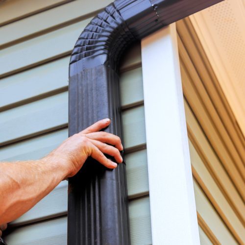 On roof, worker is installing rain gutter fitting downpipe downspout bend